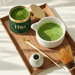 Отворена кутийка Hito Matcha върху дървен поднос с купа разбита матча, бамбукова лъжичка и бамбукова четка